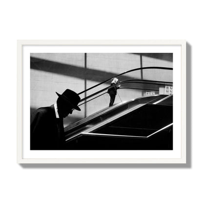 Fine art black and white street photograph of a man ascending an escalator. Archival pigment print, giant size, white frame.