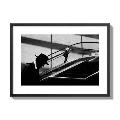 Fine art black and white street photograph of a man ascending an escalator. Archival pigment print, large size, black frame.