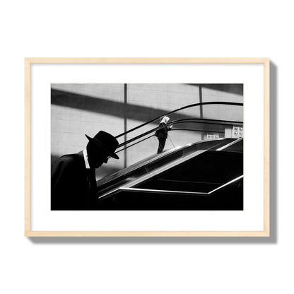 Fine art black and white street photograph of a man ascending an escalator. Archival pigment print, large size, natural wood frame.