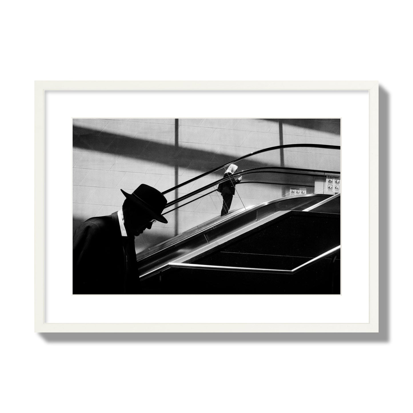 Fine art black and white street photograph of a man ascending an escalator. Archival pigment print, large size, white frame.