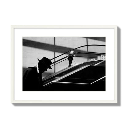 Fine art black and white street photograph of a man ascending an escalator. Archival pigment print, large size, white frame.
