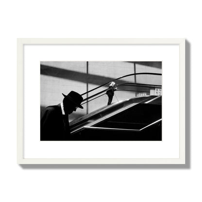 Fine art black and white street photograph of a man ascending an escalator. Archival pigment print, medium size, white frame.