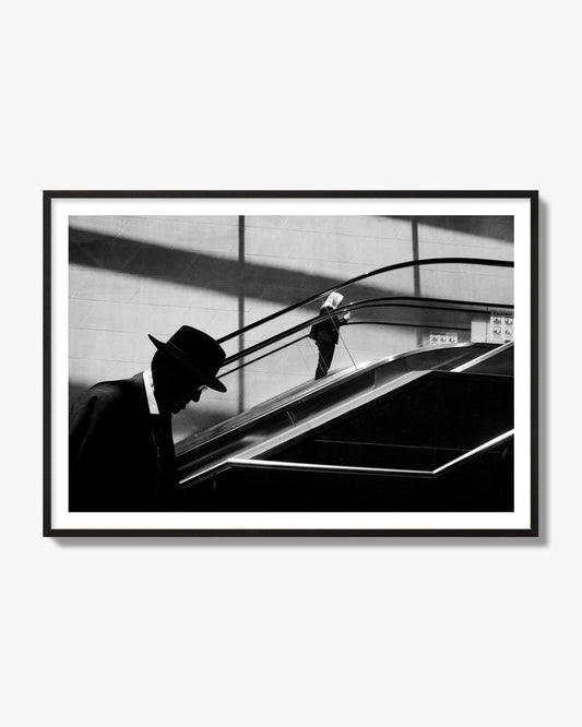 Fine art black and white street photograph of a man ascending an escalator. Archival pigment print, black frame.