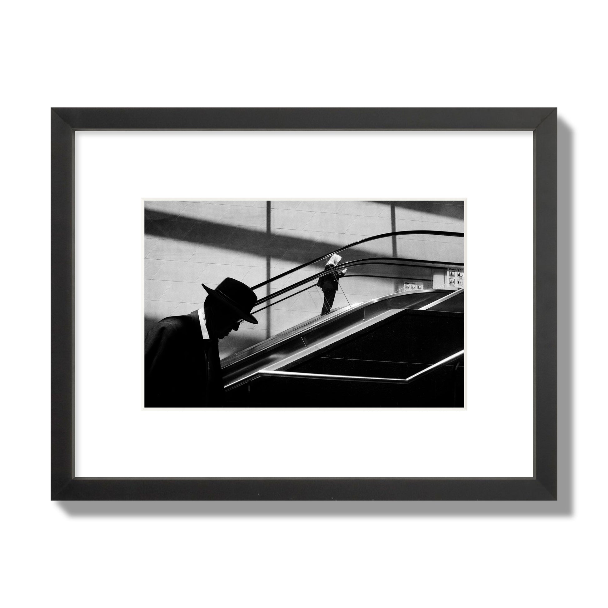 Fine art black and white street photograph of a man ascending an escalator. Archival pigment print, small size, black frame.