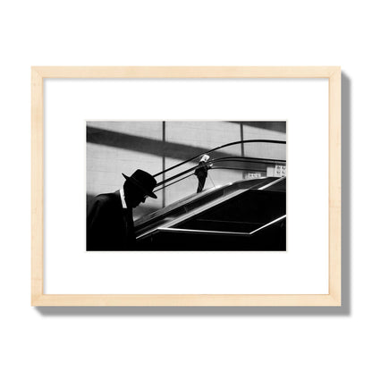 Fine art black and white street photograph of a man ascending an escalator. Archival pigment print, small size, natural wood frame.