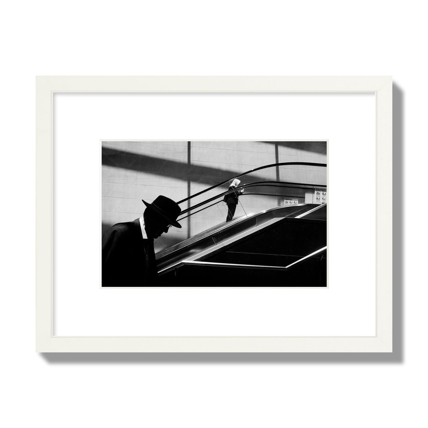 Fine art black and white street photograph of a man ascending an escalator. Archival pigment print, small size, white frame.