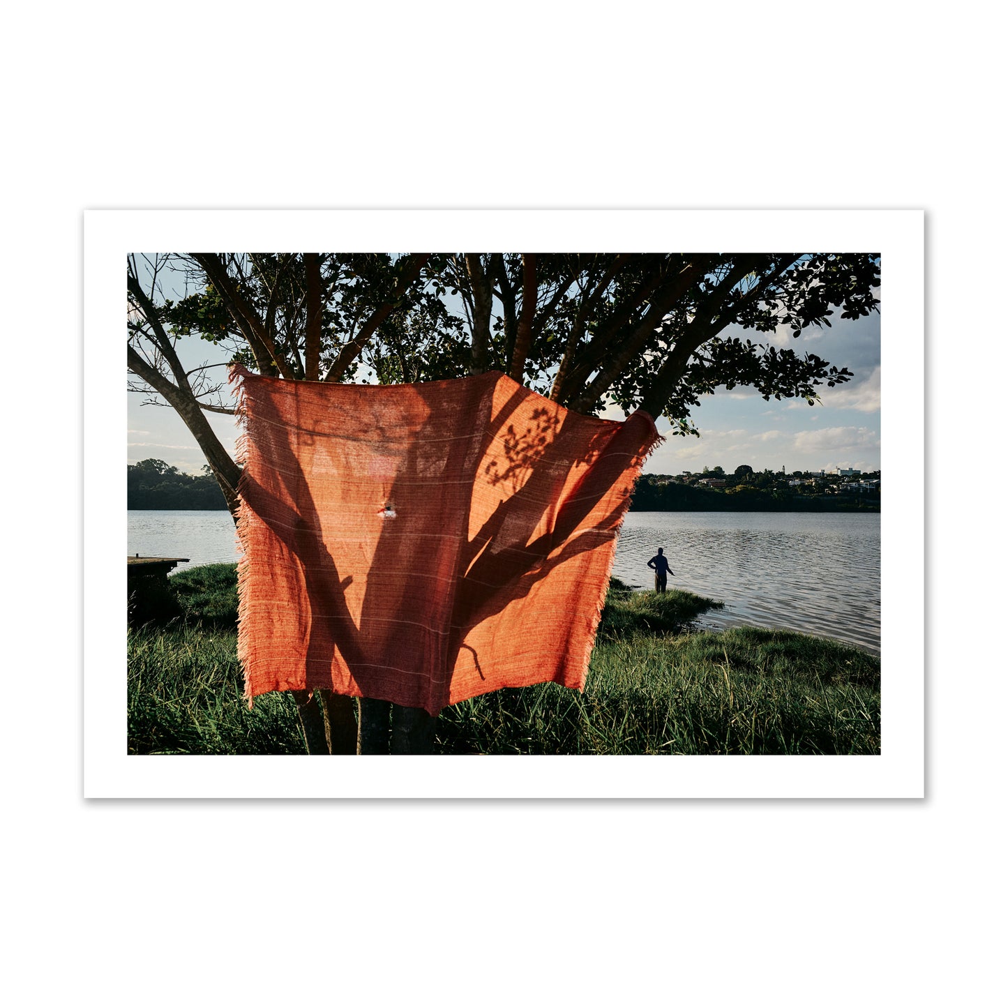 Fine art street photograph of a vibrant orange cloth hanging from a tree, with a tranquil lake and a fisherman in the background. Archival pigment print, collector size, unframed.