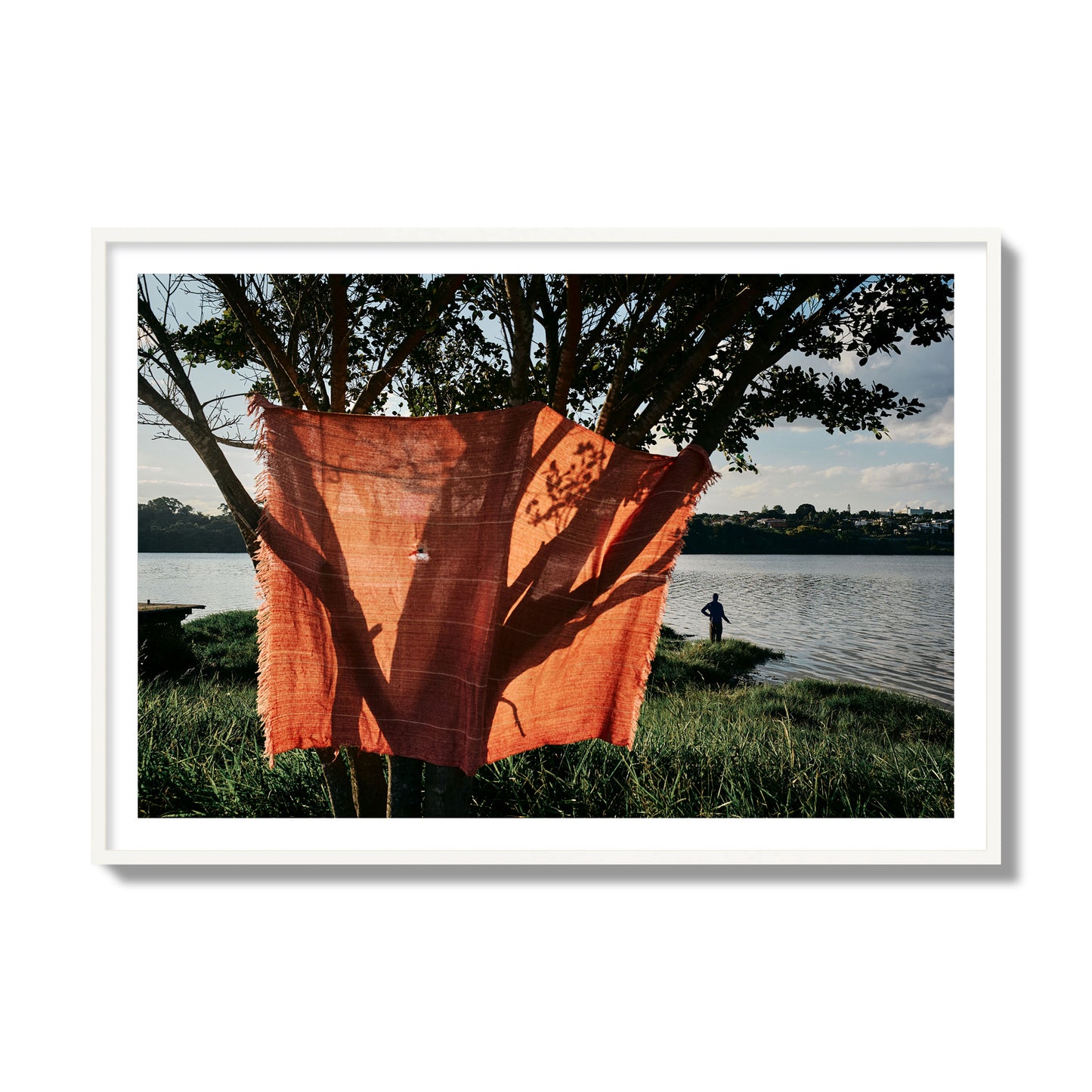 Fine art street photograph of a vibrant orange cloth hanging from a tree, with a tranquil lake and a fisherman in the background. Archival pigment print, collector size, white frame.