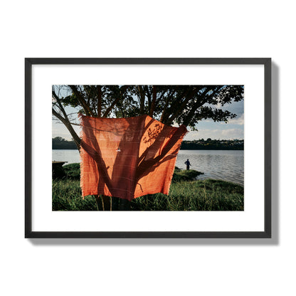 Fine art street photograph of a vibrant orange cloth hanging from a tree, with a tranquil lake and a fisherman in the background. Archival pigment print, large size, black frame.
