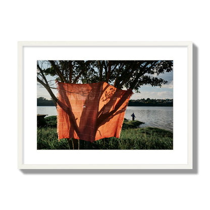 Fine art street photograph of a vibrant orange cloth hanging from a tree, with a tranquil lake and a fisherman in the background. Archival pigment print, large size, white frame.