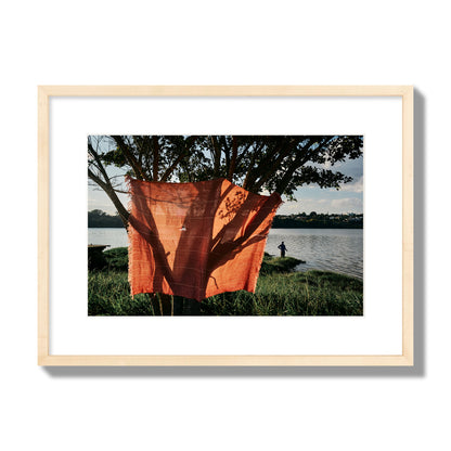 Fine art street photograph of a vibrant orange cloth hanging from a tree, with a tranquil lake and a fisherman in the background. Archival pigment print, medium size, natural wood frame.