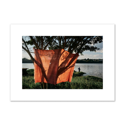 Fine art street photograph of a vibrant orange cloth hanging from a tree, with a tranquil lake and a fisherman in the background. Archival pigment print, medium size, unframed.