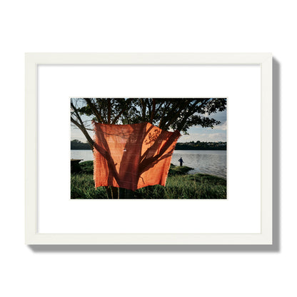 Fine art street photograph of a vibrant orange cloth hanging from a tree, with a tranquil lake and a fisherman in the background. Archival pigment print, small size, white frame.