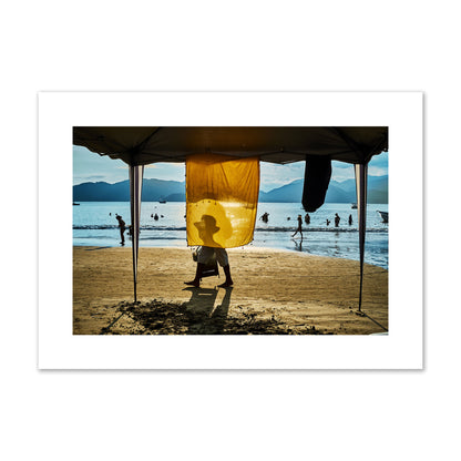 Fine art street photograph of a crowded beach scene with a vibrant yellow flap hanging from a tent, sunlight filtering through and silhouetting a man’s face. Archival pigment print, large size, unframed.