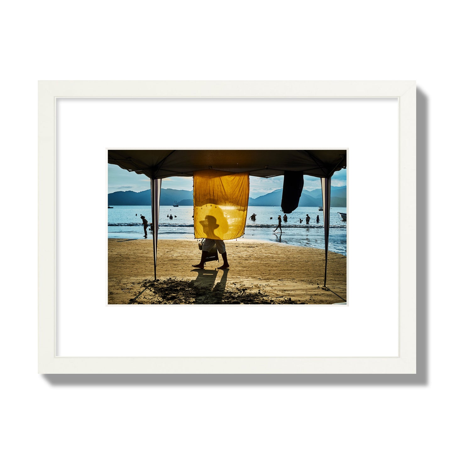 Fine art street photograph of a crowded beach scene with a vibrant yellow flap hanging from a tent, sunlight filtering through and silhouetting a man’s face. Archival pigment print, small size, white frame.