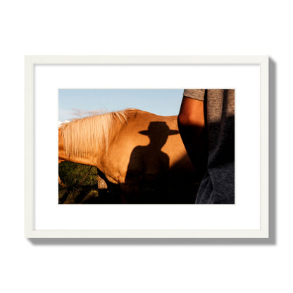 Fine art photo of a cowboy's shadow cast against the side of a horse at sunset. Archival pigment print, medium size, white frame.