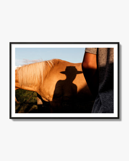 Fine art photo of a cowboy's shadow cast against the side of a horse at sunset. Archival pigment print, black frame.