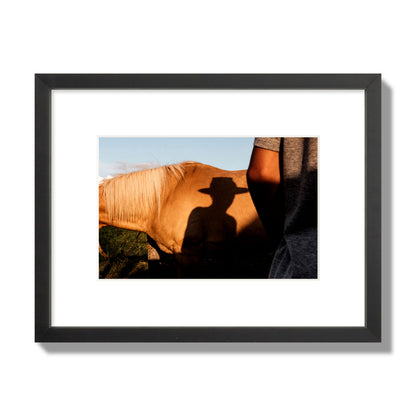 Fine art photo of a cowboy's shadow cast against the side of a horse at sunset. Archival pigment print, small size, black frame.
