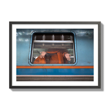 Fine art photograph of a Buddhist monk standing with open hands at a train car window. Archival pigment print, giant size, black frame.