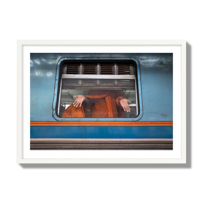 Fine art photograph of a Buddhist monk standing with open hands at a train car window. Archival pigment print, giant size, white frame.
