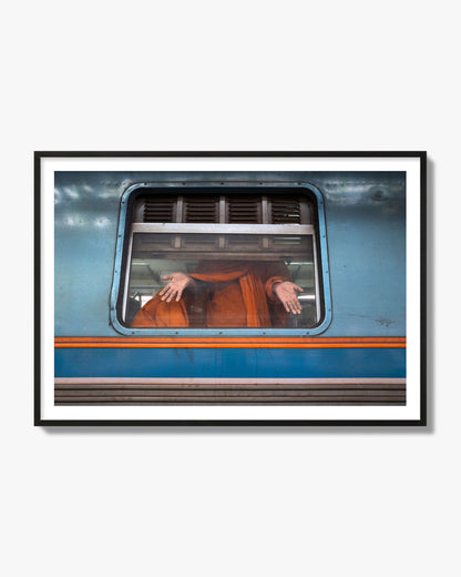 Fine art photograph of a Buddhist monk standing with open hands at a train car window. Archival pigment print, black frame.