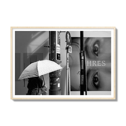 Fine art street photograph of a woman walking with an umbrella on a Tokyo street, passing beneath a billboard with two large eyes. Archival pigment print, gallery size, natural wood frame.