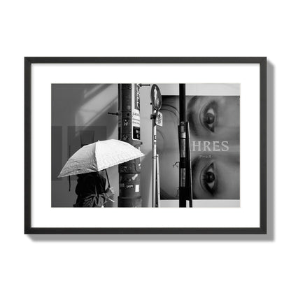 Fine art street photograph of a woman walking with an umbrella on a Tokyo street, passing beneath a billboard with two large eyes. Archival pigment print, large size, black frame.