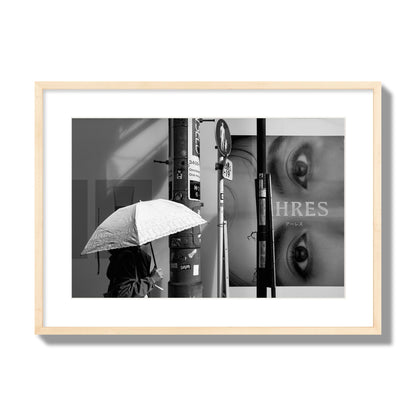 Fine art street photograph of a woman walking with an umbrella on a Tokyo street, passing beneath a billboard with two large eyes. Archival pigment print, large size, natural wood frame.