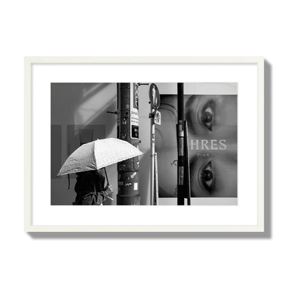 Fine art street photograph of a woman walking with an umbrella on a Tokyo street, passing beneath a billboard with two large eyes. Archival pigment print, large size, white frame.