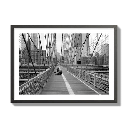 Fine art black and white photograph of a person posing on the Brooklyn Bridge with the cityscape in the background, archival pigment print, giant size, black frame