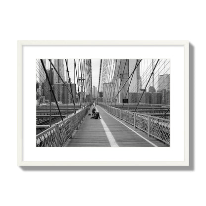 Fine art black and white photograph of a person posing on the Brooklyn Bridge with the cityscape in the background, archival pigment print, large size, white frame