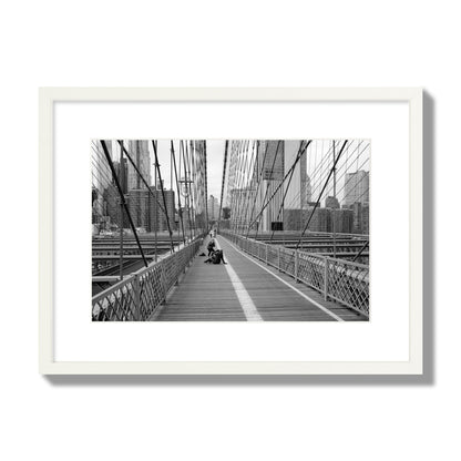 Fine art black and white photograph of a person posing on the Brooklyn Bridge with the cityscape in the background, archival pigment print, medium size, white frame