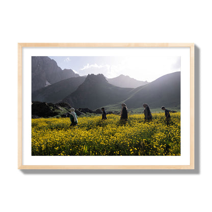 Fine art photograph of people walking through a field of yellow flowers with mountains in the background, archival pigment print, giant size, natural wood frame.