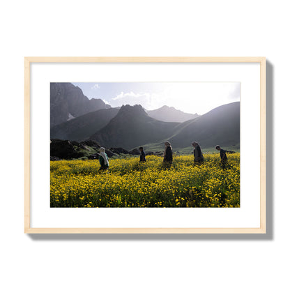 Fine art photograph of people walking through a field of yellow flowers with mountains in the background, archival pigment print, large size, natural wood frame.