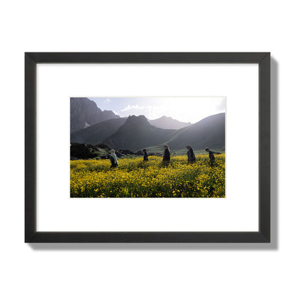 Fine art photograph of people walking through a field of yellow flowers with mountains in the background, archival pigment print, small size, black frame.