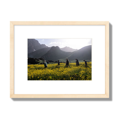 Fine art photograph of people walking through a field of yellow flowers with mountains in the background, archival pigment print, small size, natural wood frame.