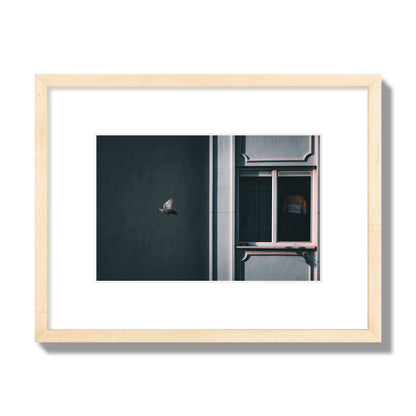 Fine art street photograph of a woman highlighted in a window on the right side of the frame, while a bird flies out of frame on the left. Archival pigment print, small size, natural wood frame.