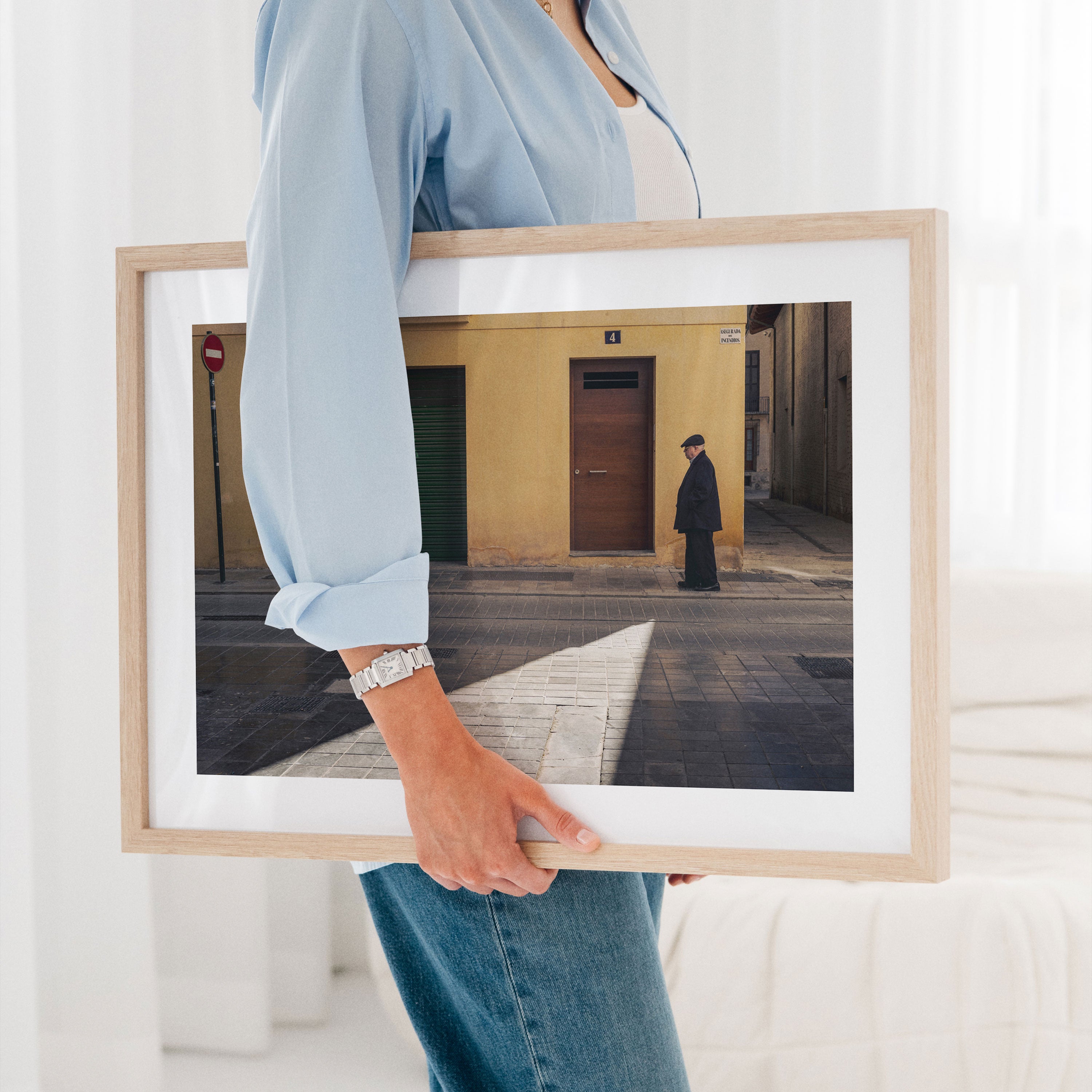 A framed fine art photograph of an elderly man walking in Valencia, Spain is tucked under a woman's arm as she walks through her living room.