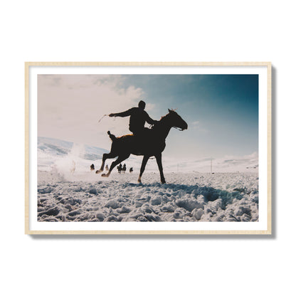 Fine art photography of a silhouetted man riding a galloping horse in a snowy field with a mountain backdrop, archival pigment print, collector size, natural wood frame.
