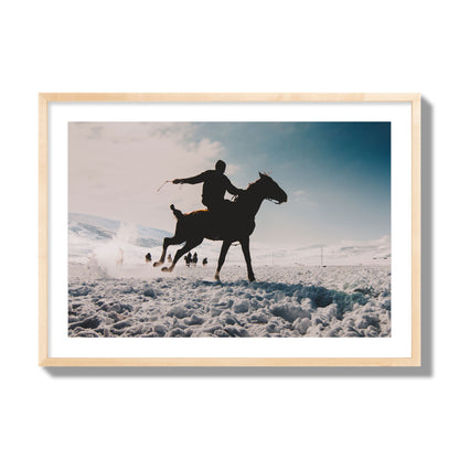 Fine art photography of a silhouetted man riding a galloping horse in a snowy field with a mountain backdrop, archival pigment print, giant size, natural wood frame.