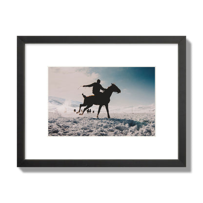 Fine art photography of a silhouetted man riding a galloping horse in a snowy field with a mountain backdrop, archival pigment print, small size, black frame.