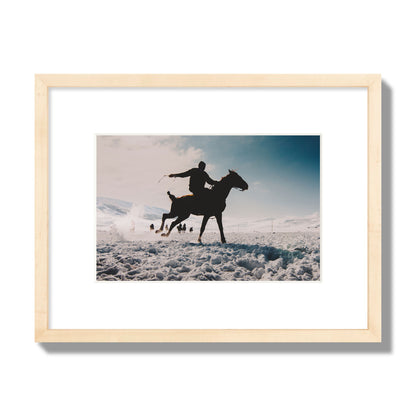 Fine art photography of a silhouetted man riding a galloping horse in a snowy field with a mountain backdrop, archival pigment print, small size, natural wood frame.