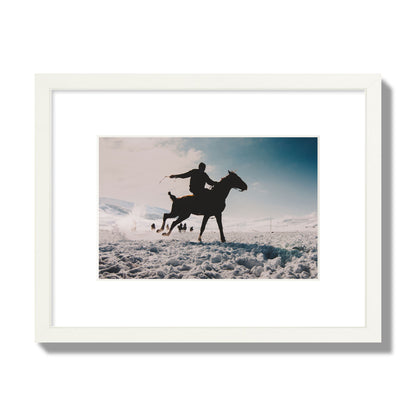 Fine art photography of a silhouetted man riding a galloping horse in a snowy field with a mountain backdrop, archival pigment print, small size, white frame.